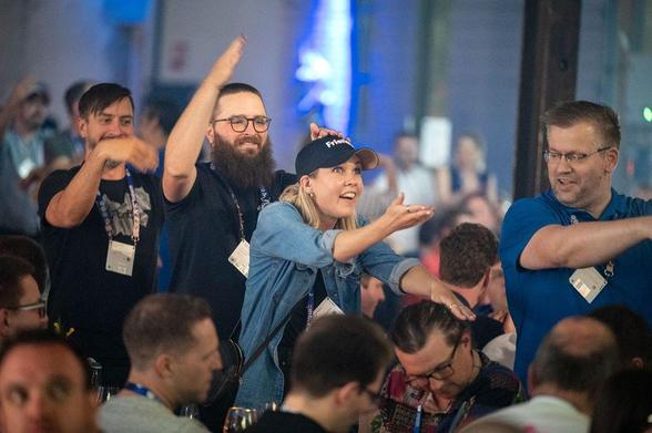 A lively group of Umbraco Codegarden attendees stand in a conga line between tables, dancing and laughing. The woman in front wears a “Friends” cap and denim shirt, leading the group with enthusiasm. The room is buzzing with energy and joyful expressions.
