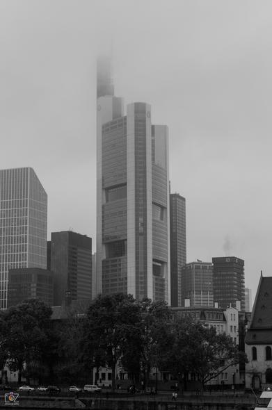 Black and white photo in portrait orientation. In the center is a skyscraper (Commerzbank Tower in Frankfurt am Main), whose upper, characteristic top sinks into the fog. The high-rise is flanked on the left and right by lower office buildings. In the foreground, obscured by trees, residential buildings and a few cars along the (invisible) banks of the river Main.