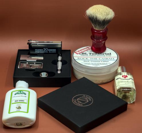 (Le Chat Mistral description)

The image showcases a collection of grooming items arranged neatly. At the centre, there's a black box containing a silver razor and additional blades. To the right, a round container of shaving soap labelled "Rock the Casbah" sits with a red-handled shaving brush on top. A bottle of cologne is positioned nearby. To the left, there's a white bottle labelled "Boat Drinks" for post-shave balm. In front, a black box with the branding "Atelier Durban" is visible. The items are set against a brown background, creating a warm, cohesive display of traditional grooming essentials.