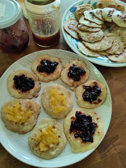 A plate with 8 neatly arranged mini pancakes, each topped with a spoonful of jam. Behind it, some jars of jam and a plate stacked with mini pancakes. The background is a wooden end grain pattern.