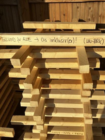 gestapelte Holzlatten in der Abendsonne, eine mit Aufschrift "Tollwood für alle - Ois inklusiv(er)! (UN-BRK)"