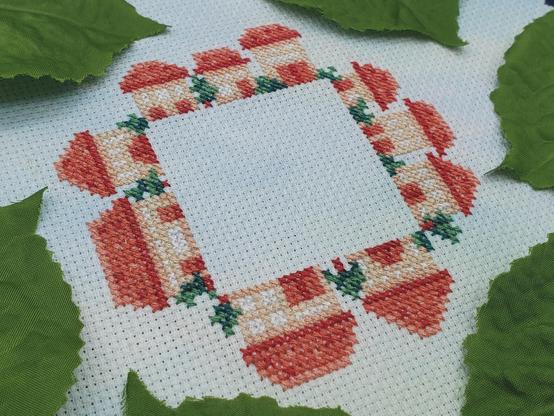 A cross stitch of tiny beige houses with red roofs and tiny trees sitting neatly along a very square perimeter.