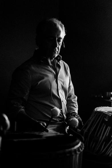 The image captures a man engrossed in playing the drum, set against a stark, dark backdrop. The foreground features parts of the drum, the drumsticks being held by the man and his cuffs. The scene exudes an atmosphere of dedicated practice or performance, the individual lost in the rhythm of his music.