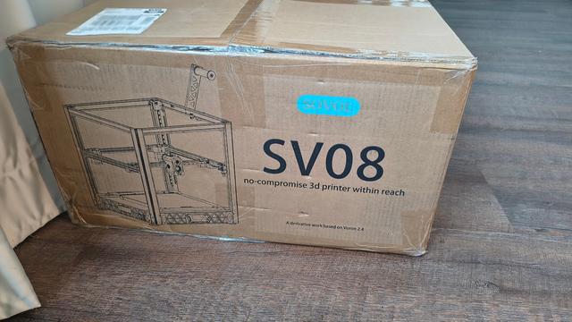 A card box box on wooden floor. The left side of the box has a drawing of a 3D printer. On the right there is the following writing:
SOVOL
SV08
no-compromise 3d printer within reach
A derivative work based on Voron 2.4