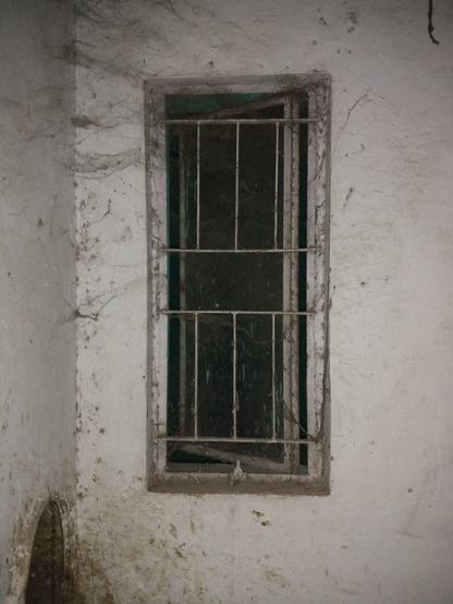 A photo of a small room with an open window surrounded by a white wall covered in dark cobwebs. To the bottom left of the image is a circular decrepit garbage chute.