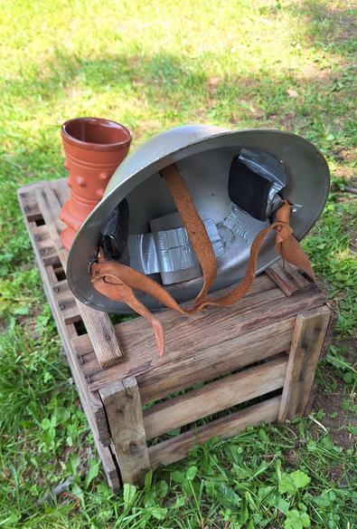 A metal helmet with leather and duct tape straps on an empty wine crate next to a clay cup on a sunny lawn.