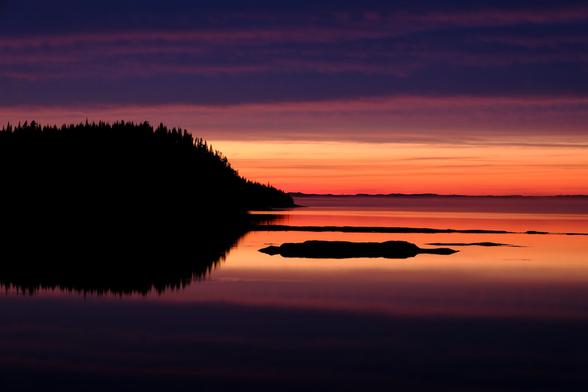 A bright band of orange and yellow colors mixed with fog paints the coast along the horizon behind a small silhouetted forest-covered island, all beneath layers of purple clouds at sunset in the Mingan archipelago in Quebec Canada.