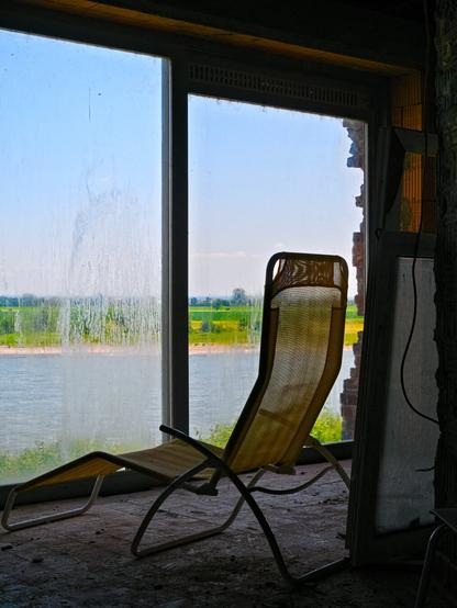 A yellowish deckchair stands in front of a large two-light window, one pane of which is blind in places. The backrest is raised so that the person sitting there has a great view of the blue waters of the Rhine, the sandy bank and the green meadows opposite. The deckchair stands on uneven ground, parts of a wall can be seen on the right and a cable hangs down.