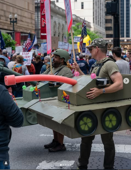 Two men, who I strongly suspect are veterans, are dressed up in papier mache tank costumes walking along in the giant No Kings crowd in Chicago. The main "gun" of each tank appears to be a pool noodle, and there are festive multicolored pinwheels to go along with that.