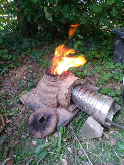 Der Lehmofen steht vor grünem Hintergrund und "spuckt" Feuer. Vorn im Ofen steckt eine  "Blechtüte" zur Belüftung.