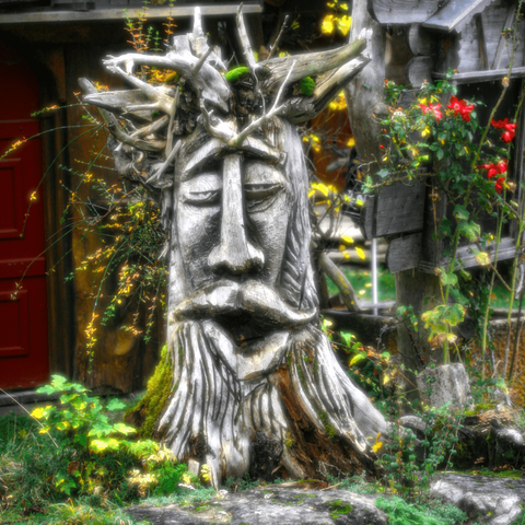 A wooden sculpture of a bearded face, the roots upwards look like chaotic hair or a crown. The eyes are half closed, the face looks deeply relaxed (or stoned). Different plants are growing around the sculpture, some with red or yellow flowers. A bloom filter effect blurs and brightens the saturated plant colors. The background consists of a brownish red door on the left and a weathered signpost on the right.