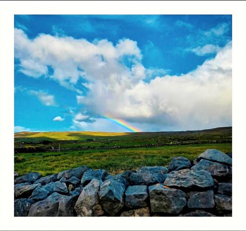 This photo is picturesque landscape, in Ireland, with a partial rainbow in the center. It is on top of a hill and it is arcing across  lots of green fields under a partly cloudy sky. The clouds are big and fluffy and take up most of the sky. The foreground is framed by a low stone wall, composed of dark, irregularly shaped, textured rocks.
