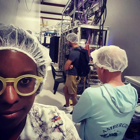 A selfie of Tiffany inside of the LIGO Hanford detector. Tiffany has dark skin and is wearing a hair net and yellow framed glasses. In the background are detector components with many wires, and two other people in hair nets.