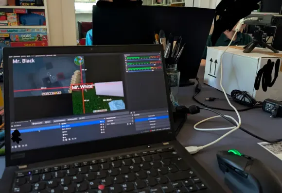 Blick auf den Redaktions-Laptop, mit OBS und beiden Game-Bildschirmen im Split-Screen. Dahinter sieht man geschwärzt die Kinder spielen. auf einer Kiste steht ein altes Zoom-Aufnahmegerät.