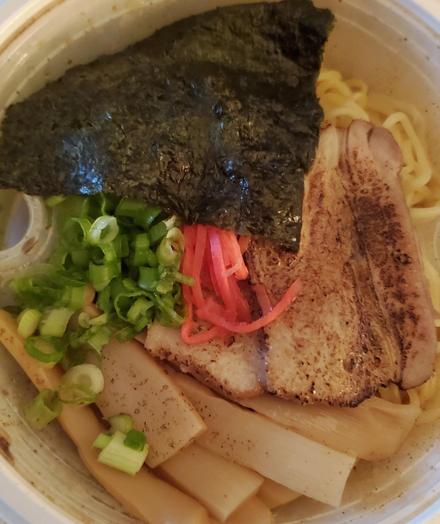 Overhead shot of a bowl of ramen. The toppings include a crinkled sheet of nori seaweed, two slices of seared pork belly, bright green sliced scallions, a small pile of pink pickled ginger, and several pale bamboo shoots. The noodles are slightly visible underneath the toppings.