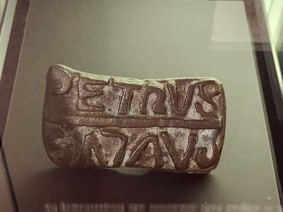 Thick, roughly rectangular bronze plate with the words “Petrus” and “Paulus” in two separate lines in thick letters. The words are positioned such that, when one is the right way up, the other is backwards and upside down.