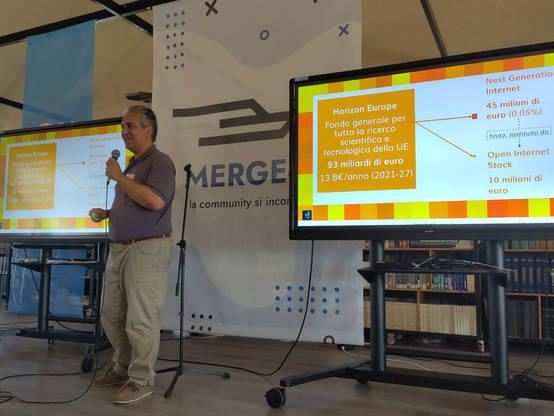 In primo piano il relatore Vittorio Bertola presenta la sessione su EuroStack, come costruire l'alternativa alle big tech. La presentazione è molto colorata e mostra i 93 miliardi di euro del fondo europeo Horizon.