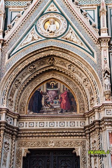 Close-up of a Duomo di Firenze portal showing multicolored marble patterns, ornate carved arches, a gilded medallion of Christ in a triangle, and a detailed mosaic of the Madonna with two saints above dark wooden doors.