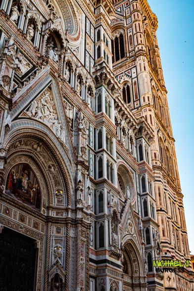 Upward view of Florence Cathedral’s richly detailed façade and Giotto’s bell tower, with multicolored marble panels, carved statues, pointed arches, and mosaics set against a clear blue sky.