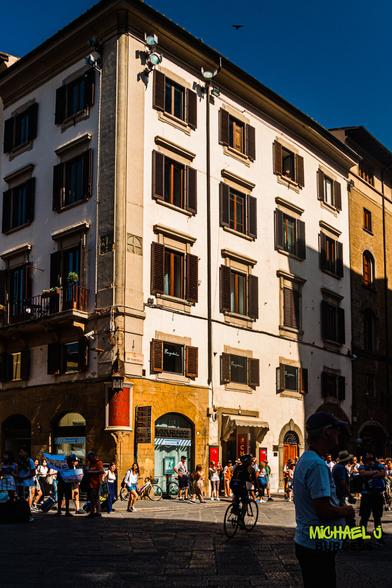Sunlit four-story white building in Florence with dark wooden shutters; pedestrians and a cyclist move through the cobblestoned street below under a deep blue sky.