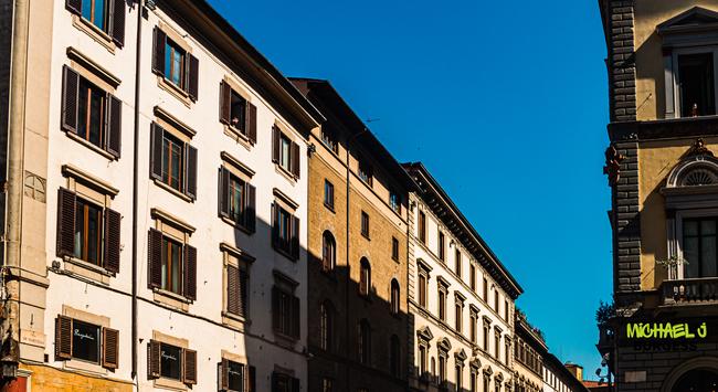 Horizontal view of a row of multi-story buildings in Florence with white and tan walls, dark wooden shutters, and deep afternoon shadows beneath a bright blue sky.