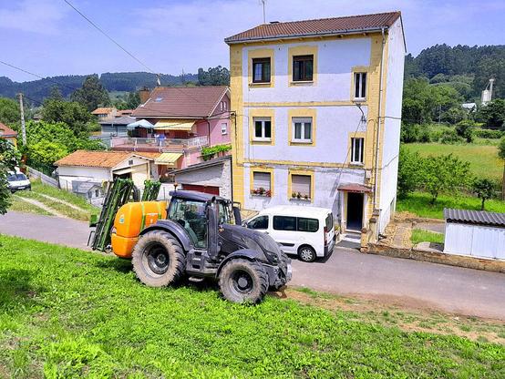 Un hermoso tractor cuál enorme escarabajo colorido, aparcado pisando la yerba delante de una humilde casa. Detrás un cuidado  chalé, unas llanas praderías y al fondo, destacando del oscuro verde de los ocalitos, la blanca torre del silo de una cooperativa agraria.