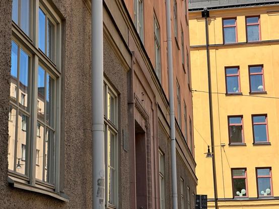 A composition with a brightly sunlit yellow apartment building, which the camera is looking towards, occupying the right ⅓, and a shadowed row of buildings to the left. Immediately in front of the camera, and occupying the leftmost ⅓, is a window, reflecting another brightly lit building, this time off to the side