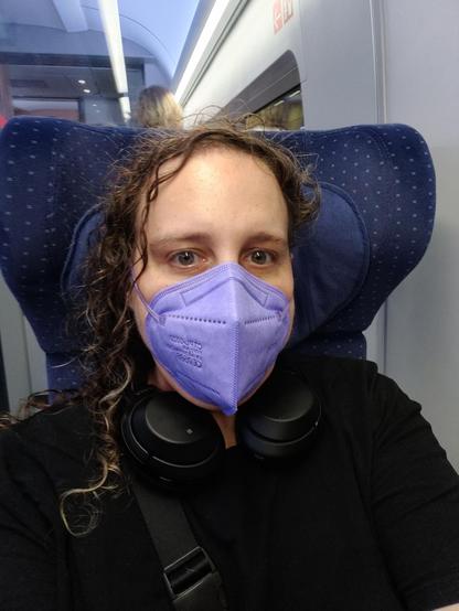Mirah sitting in an ICE wearing a black dress, black headphones around her neck, a black bag, and a lavender mask