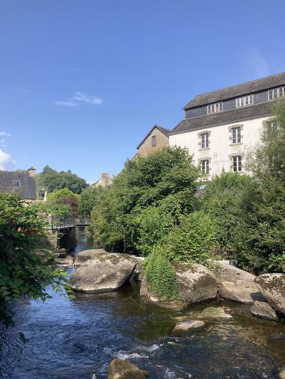 Vue d'une rivière avec de gros rochers, des arbres et une grande maison en haut à droite ; un pont et d'autres maisons plus loin à gauche