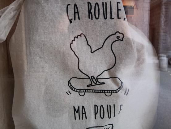 Foto einer schwarz bedruckten Baumwolltasche hinter einem Schaufenster. Auf der Tasche ist, in einfachen Strichen, ein Skateboard fahrendes Huhn und der Spruch: Ca roule, ma Poule. Auf der rechten Seite sind Spiegelungen zu erkennen, ein Poller zum Beispiel.