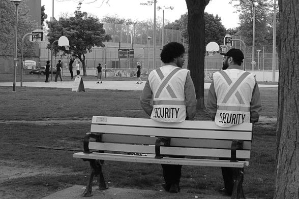 At right, backs to the camera, two bearded men in hi-vis security vests lean against a bench. They are deep in conversation. In the upper right background, a group of teens play basketball at one of the open air courts