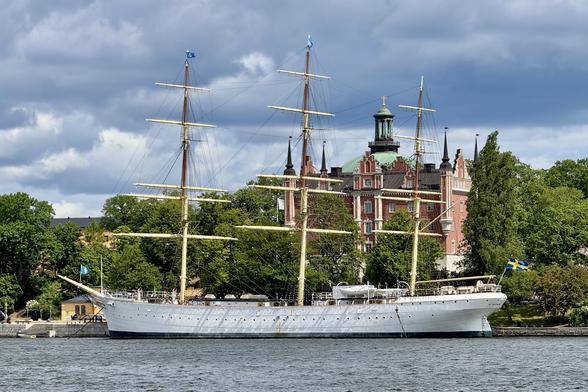 A tall ship with multiple masts is anchored in a body of water, surrounded by greenery and a historic building in the background. The ship displays a Swedish flag, and the sky is partly cloudy.