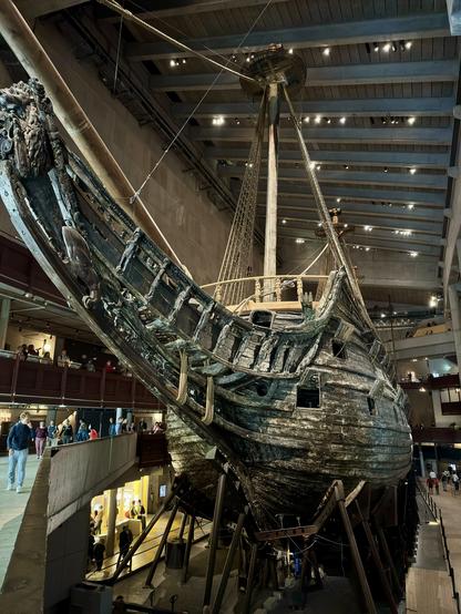 The image shows a large, historic ship displayed in a museum. The ship, partially submerged, has an ornate wooden structure with detailed carvings and ropes. Surrounding the vessel are visitors and architectural elements of the museum, including viewing areas and walkways.