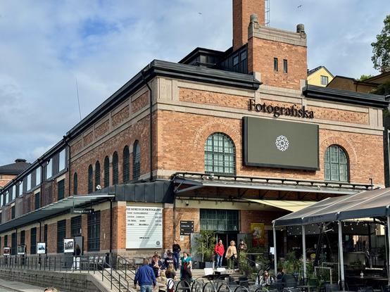 The image shows the exterior of Fotografiska, a photography museum, featuring a large brick building with arched windows and a prominent sign displaying the museum's name. People are gathered outside, and there are outdoor seating areas and a café present.