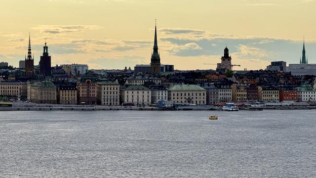 A panoramic view of a city skyline at sunset, featuring historic buildings with distinctive spires and a harbor with a small boat in the foreground. The scene highlights a mix of architectural styles and a calm water surface reflecting the colorful sky.