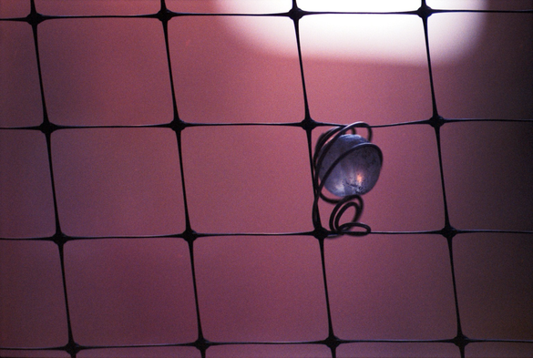 A small glass sphere, catching the sun but dirty from years outdoors, is mounted in a spiral of wire that is caught in the grid of balcony pigeon netting. The background is a dusky rose with a bright splash in the upper right - actually sunlight and shadow of an out-of-focus brick wall.