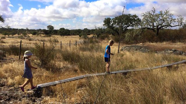 Two children walk along a narrow wooden footbridge through dry grassland at Bubulcus & Bolotas. One wears a sunhat and dress, the other carries a stick. Sparse trees and a wire fence stretch across the warm, mediterranean landscape under a partly cloudy sky.
