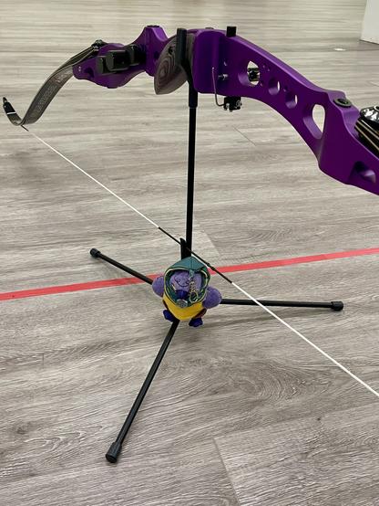 A purple recurve bow rests on a black bow stand. The bow stand is adorned with a small plush bird-like character, Kiri, from the Dungeons and Dragons streaming show Critical Role