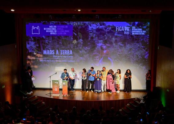 A imagem mostra uma cerimônia de premiação no FICA (Festival Internacional de Cinema e Vídeo Ambiental). O telão exibe o título e os detalhes do filme “Mãos à Terra”, bem como os logotipos do festival. A equipe está recebendo um prêmio, enquanto o público se senta no teatro escuro abaixo.