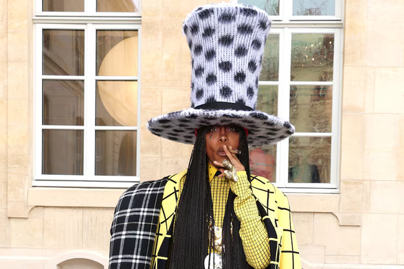 Erykah Badu wears a dramatic tall white/grey top hat with black polka dots, and has long black braids cascading down. Their outfit consists of a bright yellow suit with black grid lines, paired with a black and white plaid coat or cape draped over one shoulder.