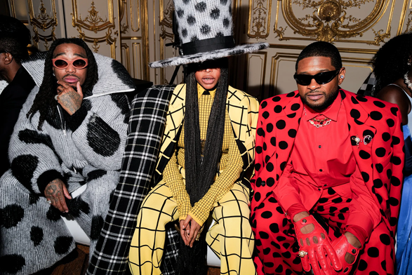 Three people sit together in an ornate room with gold baroque-style wall decorations and molding. On the left, a Black man with long dark hair wears pink sunglasses and a black and white patterned oversized sweater/jumpsuit. In the center, Erykah Badu wears a distinctive tall white hat with black polka dots, long braids, and a yellow outfit with black grid lines. On the right, Usher wears dark sunglasses and a bright red outfit covered in black polka dots, holding red gloves..