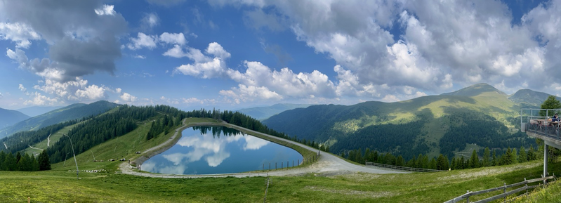 Bergpanorama mit dem Speicherteich in dem sich im Wasser die Wolken spiegeln und mit grünen Hügeln rundum