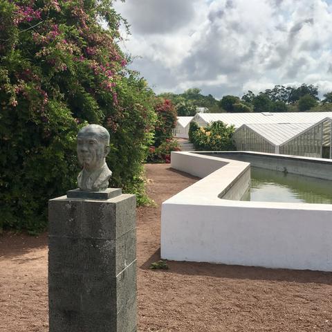 A bronze bust stands on a plinth in front of the white-walled water reservoir and, further back, greenhouses for the cultivation of pineapples.