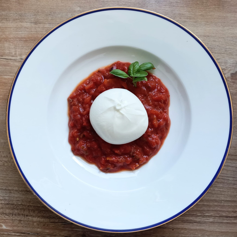 Auf einem Teller das rote Tomatenragout mit einer Kugel Burrata und einem Zweig Basilikum