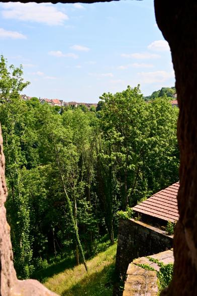 Ein Blick durch alte Steinmauern zeigt eine üppige Landschaft voll grüner Bäume und einen Teil der Außenmauer, auf einer Hochebene sind Häuser in der Ferne zu erkennen, unter einem strahlend blauen Himmel mit verstreuten Wolken.