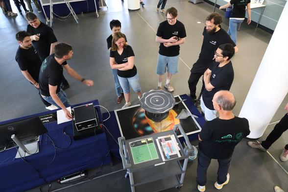 A view from above onto an experiment, surrounded by people looking at it, turning knobs, and pointing. The center piece of the experiment is a large flat dark plate with some amount of sand on it that collects in the node lines of the vibrations of the plate. Those vibrations are caused by a massive speaker sitting directly under the plate.