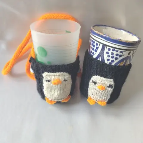 Un porte-gobelet noir avec une face de manchot crème et une lanière orange, il contient un gobelet en plastique. À sa droite (sur la photo) un autre porte-gobelet sans la lanière. Dedans : un gobelet en faïence tunisienne.