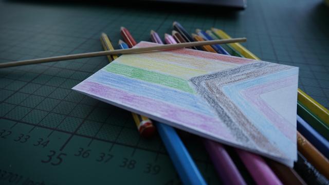 In the picture is a folded piece of paper, coloured by pen to be a pride flag, next to it is a little wooden stick, which will become the flagpole. Beneath the paper, there lay the pens I've used for colouring. And they lay on my cutting mat on my desk.