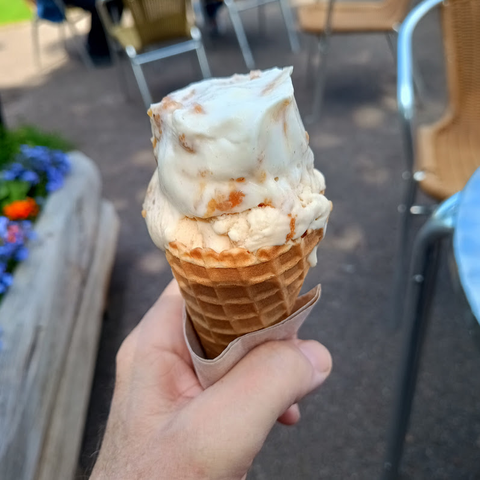 A big mound of white ice cream with orange/brown bits in it sits in a wafer cone help in my hand. Out of focus outdoor cafe furniture in the background.
