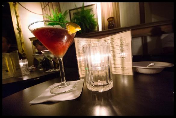A strawberry lemon martini sits on a bar table... softly illuminated by a candle. A martini menu behind the candle reflects the light and promises much deliciousness.